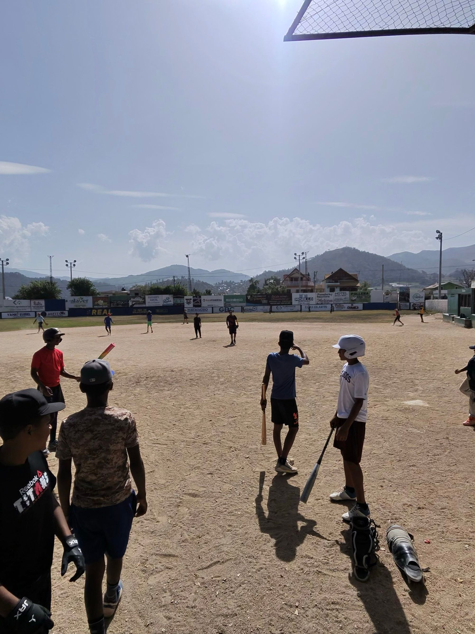 Niños con bates y un casco en el infield de tierra en Constanza, con las montañas detrás de la cerca