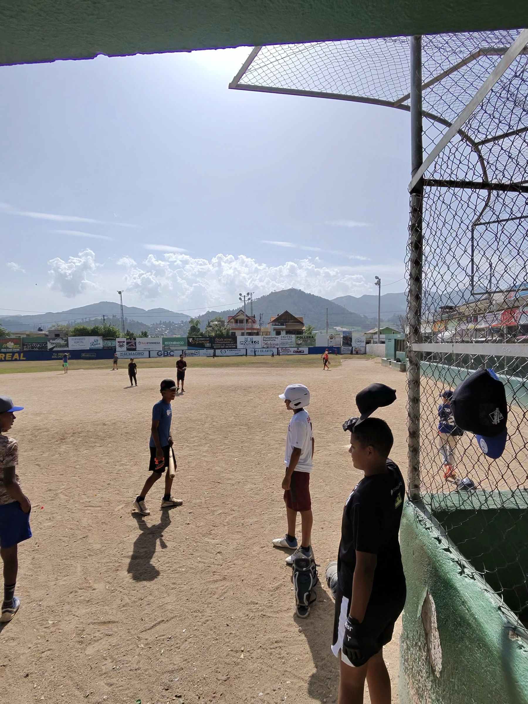 Vista desde el dugout a través de la cerca: niños esperando su turno al bate, gorras colgadas de la malla